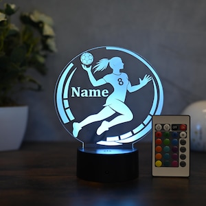 Puede incluir: Una silueta blanca de una persona jugando al balonmano con una pelota en la mano, con la palabra "Name" y el número "8" en el diseño. El diseño está sobre una base redonda de acrílico transparente con una base negra y un mando a distancia con luces LED de cambio de color.