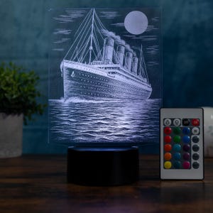 Peut inclure: Veilleuse LED rectangulaire avec une illustration détaillée d'un navire naviguant sur l'eau sous une pleine lune. La lumière a une base noire et comprend une télécommande avec des options de couleurs.
