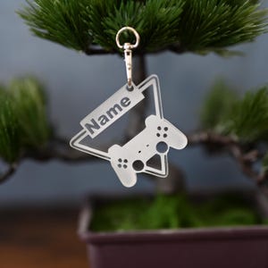 Könnte beinhalten: Ein transparenter Schlüsselanhänger aus Acryl mit einem Videospiel-Controller-Design und Platz für einen Namen. Der Name "Name" ist auf den Schlüsselanhänger gedruckt.
