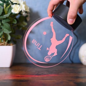 May include: A pink acrylic night light with a silhouette of a tennis player and the name "Tina" inscribed on it. The light is illuminated and casting a red glow on the surface below.