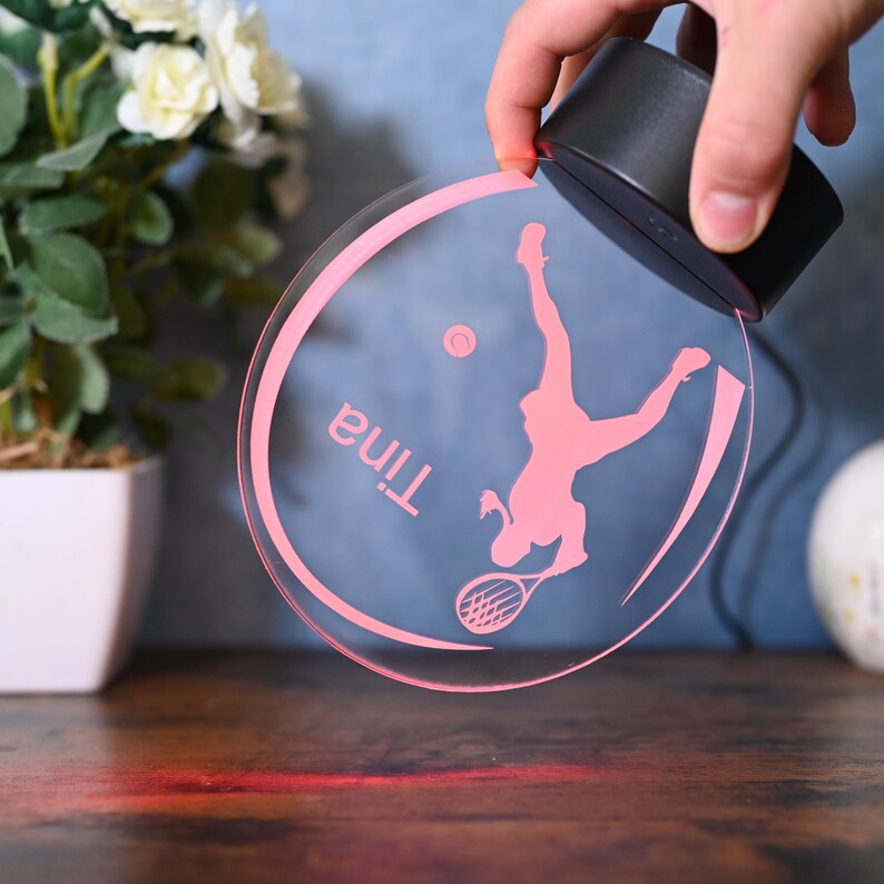 May include: A pink acrylic night light with a silhouette of a tennis player and the name "Tina" inscribed on it. The light is illuminated and casting a red glow on the surface below.