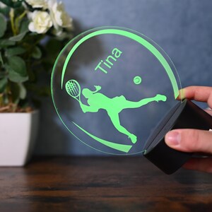 May include: A green illuminated acrylic sign with a silhouette of a tennis player and a tennis ball. The sign has the name "Tina" written on it.
