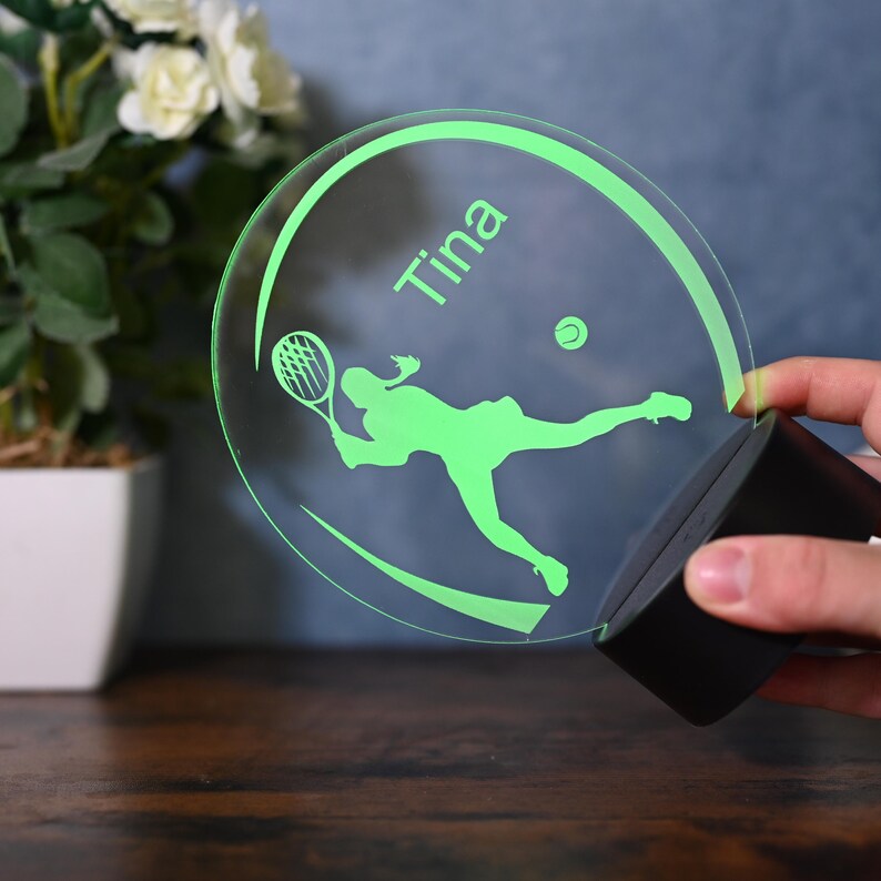 May include: A green illuminated acrylic sign with a silhouette of a tennis player and a tennis ball. The sign has the name "Tina" written on it.