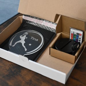 May include: A white box with a clear acrylic plaque featuring a silhouette of a tennis player and the name "Tina". The box also contains a black base with a remote control for changing the color of the light.