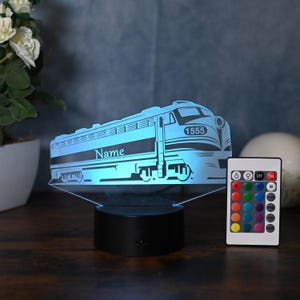 May include: A blue 3D printed night light shaped like a train with the number 1555 on the side. The train has the word "Name" printed on the side. The night light is on a black base and comes with a remote control.