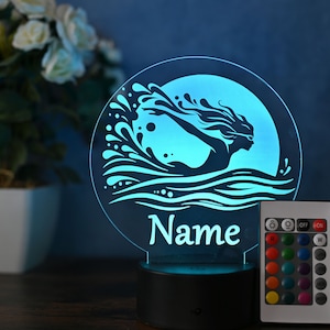 Può includere: Una luce notturna stampata in 3D blu e bianca con una silhouette di una donna che nuota tra le onde. La luce ha un telecomando con più opzioni di colore e il testo "Nome" è stampato sulla base.