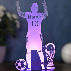 May include: A purple illuminated acrylic silhouette of a soccer player raising their arms in victory. The player is wearing a jersey with the number 10 and the word "Name" on it. The silhouette is standing on a base with a soccer ball and a trophy.