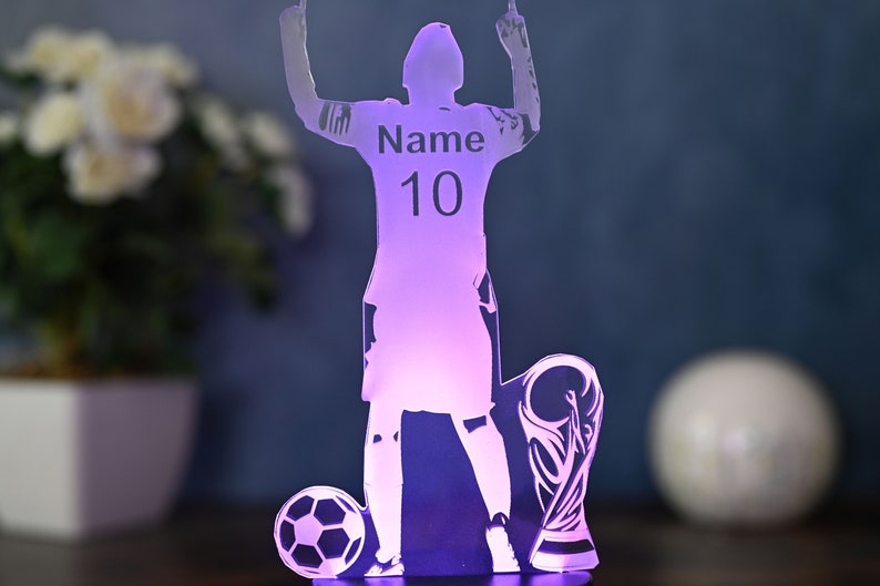 May include: A purple illuminated acrylic silhouette of a soccer player raising their arms in victory. The player is wearing a jersey with the number 10 and the word "Name" on it. The silhouette is standing on a base with a soccer ball and a trophy.