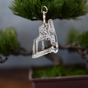 Könnte beinhalten: Transparenter Acryl-Schlüsselanhänger mit einem goldfarbenen Verschluss. Der Schlüsselanhänger hat die Form eines Maschinenteils, in das das Wort "Name" eingeätzt ist. Der Schlüsselanhänger hängt an einem kleinen grünen Ast.