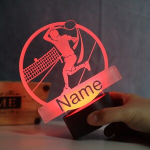 Op de afbeelding: Rood acryl nachtlampje met een silhouet van een tennisspeler in een cirkelvormig frame. Het lampje heeft een voet en een apart stuk met het woord "Naam" erop.
