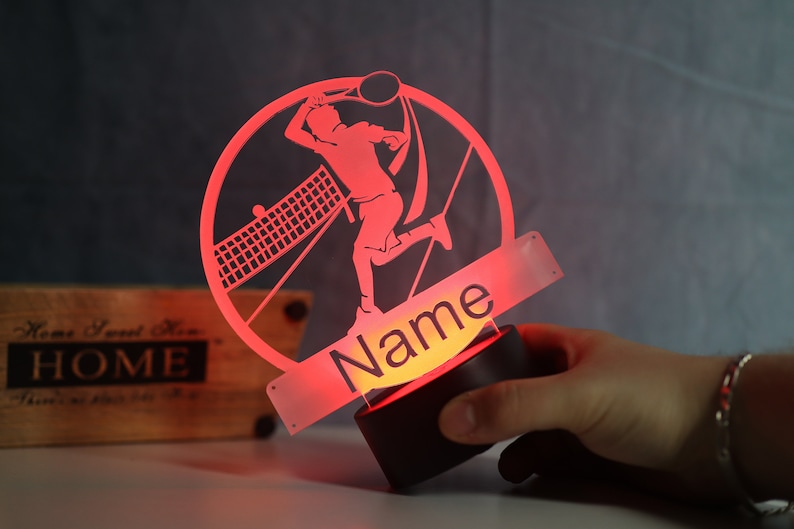 Op de afbeelding: Rood acryl nachtlampje met een silhouet van een tennisspeler in een cirkelvormig frame. Het lampje heeft een voet en een apart stuk met het woord "Naam" erop.