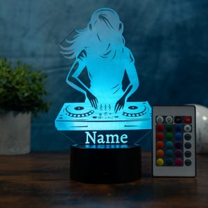 Op de afbeelding: Blauwe LED-lamp met een DJ-silhouet met koptelefoon en handen op een draaitafel, met het woord "Name" aan de basis. Inclusief een afstandsbediening. De lamp staat op een zwarte voet.