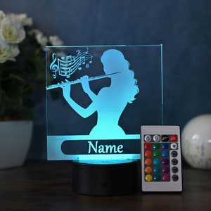 Peut inclure: Une veilleuse en verre bleu avec une silhouette d'une personne jouant de la flûte. La lumière a une plaque de nom personnalisée qui dit "Nom" et est livrée avec une télécommande pour changer la couleur de la lumière.