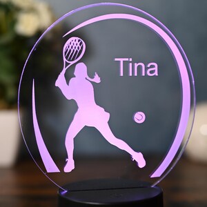 May include: A purple silhouette of a tennis player with a racket and a tennis ball, with the name "Tina" inscribed on a clear acrylic night light.