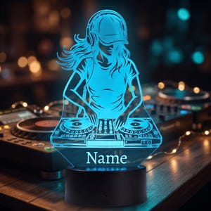 Op de afbeelding: Een oplichtende cyaan LED-acryllamp met het silhouet van een DJ die een koptelefoon en pet draagt terwijl deze een mengpaneel bedient. De voet van de lamp is zwart en cilindervormig, met het woord Name in het wit op het acryloppervlak.