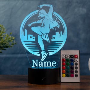 May include: A blue LED night light featuring a dancer silhouette within a circular design, with the word "Name" below. Includes a remote control for color adjustments. The base is black.