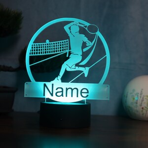 Op de afbeelding: Een turquoise nachtlampje met een silhouet van een tennisspeler in een cirkel. De basis van het lampje is zwart en de naam "Name" staat op een rechthoekig, doorzichtig paneel onder de cirkel.