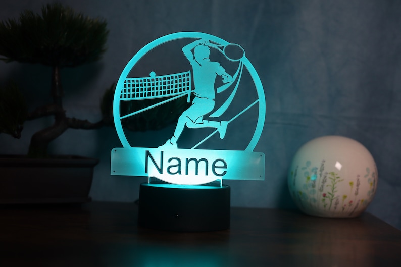 Op de afbeelding: Een turquoise nachtlampje met een silhouet van een tennisspeler in een cirkel. De basis van het lampje is zwart en de naam "Name" staat op een rechthoekig, doorzichtig paneel onder de cirkel.