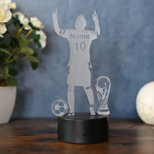 May include: A clear acrylic night light shaped like a soccer player raising their arms in victory. The player is wearing a jersey with the number 10 and the word "Name" on the back. The player is standing next to a soccer ball and a trophy. The night light has a black base.