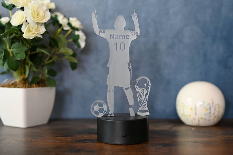 May include: A clear acrylic night light shaped like a soccer player raising their arms in victory. The player is wearing a jersey with the number 10 and the word "Name" on the back. The player is standing next to a soccer ball and a trophy. The night light has a black base.
