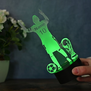 May include: Green acrylic 3D night light featuring a silhouette of a soccer player celebrating a goal. The player is holding a soccer ball and a trophy. The light has a black base and the player's jersey has the text "Name 10" on it.