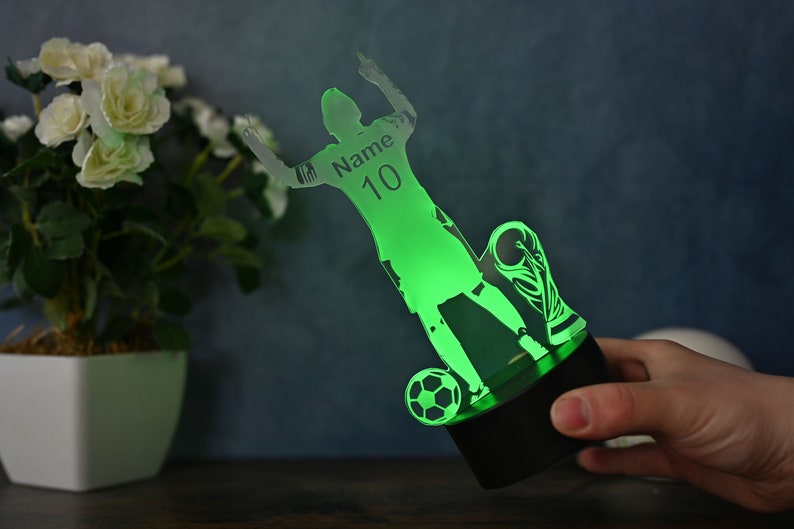 May include: Green acrylic 3D night light featuring a silhouette of a soccer player celebrating a goal. The player is holding a soccer ball and a trophy. The light has a black base and the player's jersey has the text "Name 10" on it.