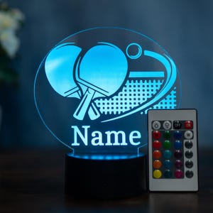 Könnte beinhalten: Eine blaue LED-Tischtennislampe mit einem Schläger- und Ball-Design. Die Lampe trägt das Wort "Name" in Weiß und wird mit einer Fernbedienung zur Farbeinstellung geliefert. Die Lampe steht auf einem schwarzen Sockel.