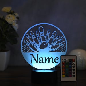 Puede incluir: Una luz nocturna impresa en 3D blanca con un diseño de bola y bolos de bolos. La luz está sobre una base negra y viene con un control remoto que ofrece múltiples opciones de color. El texto "Nombre" se muestra en la luz.