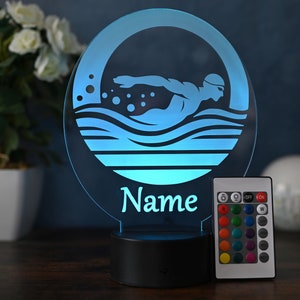 Könnte beinhalten: Ein blaues und weißes 3D-gedrucktes Nachtlicht mit einer Schwimmer-Silhouette und Wellen. Das Licht hat einen runden Sockel und eine Fernbedienung mit mehreren Farboptionen. Das Licht ist mit dem Namen "Name" personalisiert.