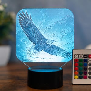 Puede incluir: Lámpara LED azul con una imagen grabada de un águila en vuelo. La lámpara tiene una base negra y un mando a distancia con múltiples opciones de color. El diseño del águila es detallado e iluminado.