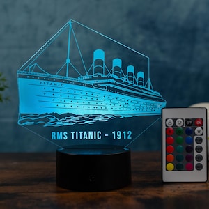 Pode incluir: Uma luz noturna LED azul com o RMS Titanic. O navio é iluminado com um brilho azul, e a base é preta. O texto "RMS TITANIC - 1912" é exibido abaixo do navio. Um controle remoto está incluído.