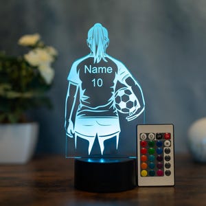 Könnte beinhalten: LED-Nachtlicht mit Fußballmotiv, das einen Spieler mit Ball zeigt, mit dem Text "Name 10" auf dem Trikot. Es hat einen schwarzen Sockel und eine Fernbedienung mit verschiedenen Farboptionen.