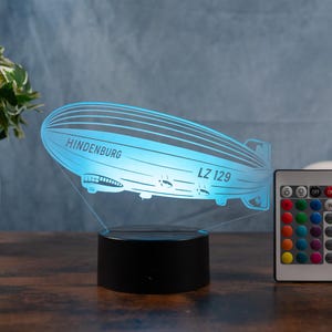 Puede incluir: Luz nocturna LED azul con forma de dirigible Hindenburg, con el texto "Hindenburg LZ 129". La luz se encuentra sobre una base negra e incluye un mando a distancia para ajustar el color.