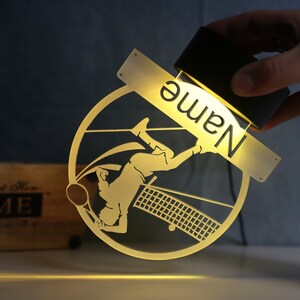 Op de afbeelding: Een gouden metalen plaquette met een silhouet van een tennisspeler en een tennisnet. De plaquette heeft ruimte voor een naam, die wordt verlicht door een lichtbron. De naam "Name" is zichtbaar op de plaquette.