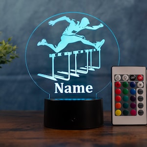 Puede incluir: Una luz nocturna LED azul con un corredor de vallas saltando vallas. El diseño está dentro de un marco circular, con la palabra "Name" debajo. Un mando a distancia está al lado de la luz.