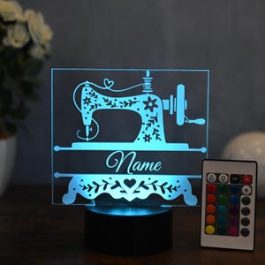May include: A blue and white personalised night light with a floral design of a sewing machine. The light has a remote control with multiple colour options. The name "Name" is printed on the light.