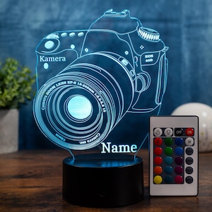 Può includere: Una lampada a LED blu a forma di fotocamera con le parole "Kamera" e "Name" incise nell'acrilico. La lampada presenta un design con obiettivo zoom e include un telecomando per la regolazione del colore. La lampada è alta circa 18 cm.