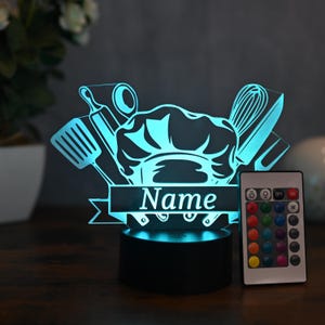 May include: A blue and white 3D printed night light shaped like a chef's hat with kitchen utensils. The light is on a black base and has a remote control with colour changing options. The light is personalised with the name "Name".