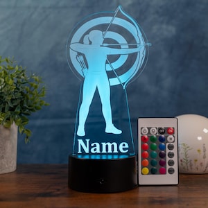 Op de afbeelding: Blauwe LED-lamp met een boogschutter die op een doelwit mikt. De lamp heeft een zwarte basis en een afstandsbediening voor kleurveranderingen. Het woord "Name" wordt aan de voet van de boogschutter weergegeven.