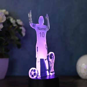 May include: A clear acrylic 3D night light shaped like a soccer player holding a trophy. The player is raising their arms in celebration. The light is illuminated in a purple hue. The player's jersey has the text "Name 10" printed on it.