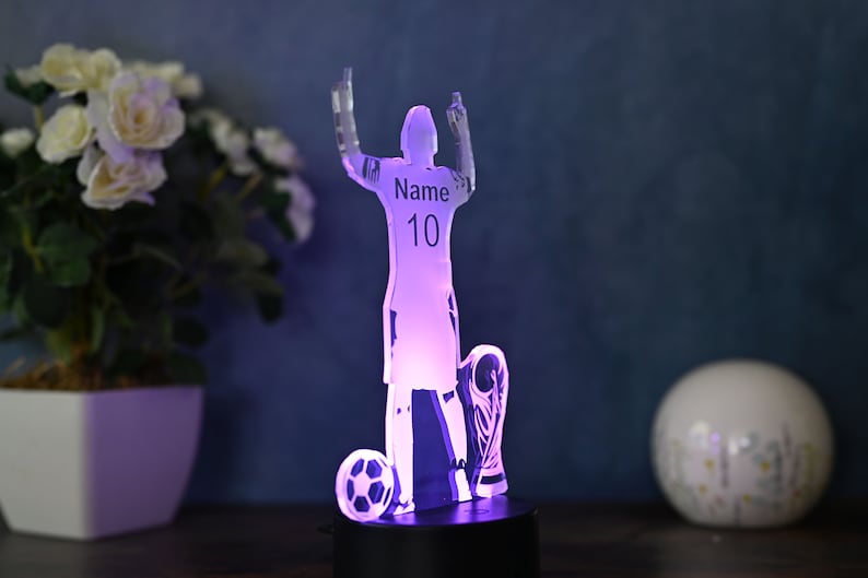 May include: A clear acrylic 3D night light shaped like a soccer player holding a trophy. The player is raising their arms in celebration. The light is illuminated in a purple hue. The player's jersey has the text "Name 10" printed on it.