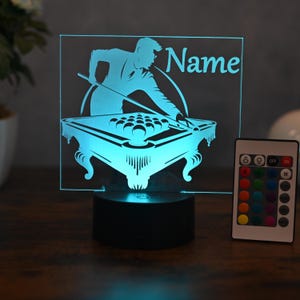 Może przedstawiać: Turkusowa lampa LED przedstawiająca gracza w bilard celującego w stół bilardowy z kulami. Lampa ma napis "Name" i jest dostarczana z pilotem do regulacji koloru. Lampa stoi na czarnej podstawie.