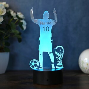 May include: A blue 3D printed acrylic night light shaped like a soccer player raising their arms in victory. The player is holding a soccer ball and a trophy. The player's jersey has the text "Name 10" printed on it.