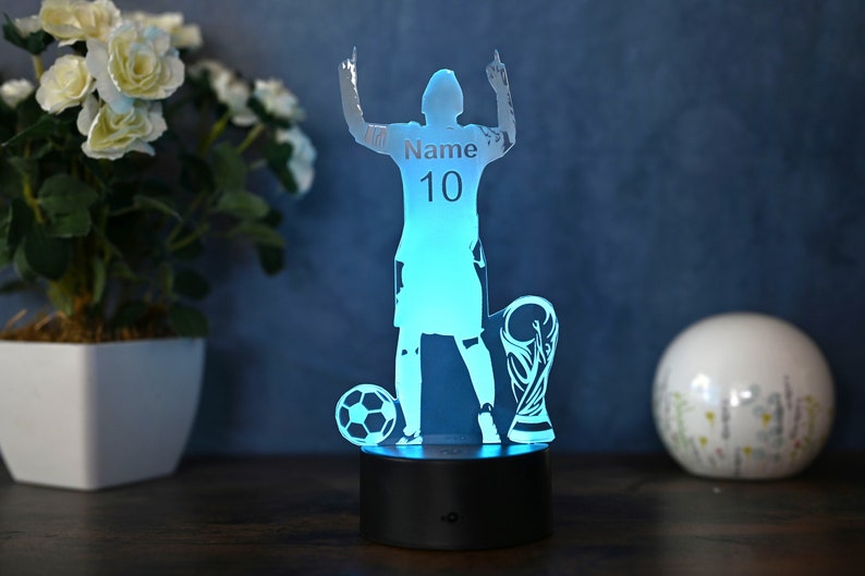 May include: A blue 3D printed acrylic night light shaped like a soccer player raising their arms in victory. The player is holding a soccer ball and a trophy. The player's jersey has the text "Name 10" printed on it.