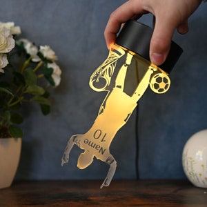 May include: A gold-colored silhouette of a soccer player kicking a ball, with the number "10" and the word "Name" printed on the player's jersey. The silhouette is illuminated from behind, creating a glowing effect. The silhouette is attached to a black base.
