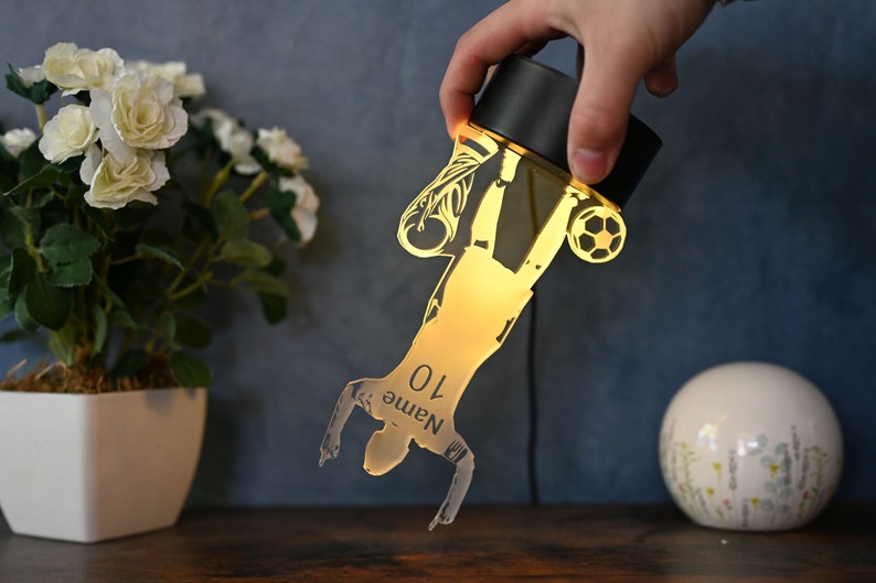 May include: A gold-colored silhouette of a soccer player kicking a ball, with the number "10" and the word "Name" printed on the player's jersey. The silhouette is illuminated from behind, creating a glowing effect. The silhouette is attached to a black base.
