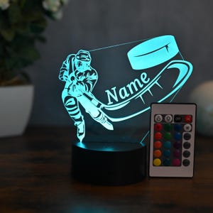 May include: A 3D printed hockey player night light with a puck and stick. The light is on a black base and has a remote control with multiple color options. The light is personalized with the name "Name".
