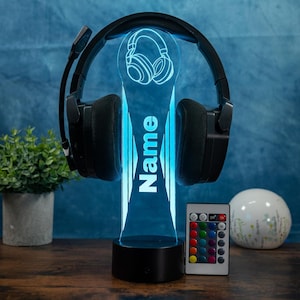 Op de afbeelding: Een zwarte gamingheadset rust op een blauwe LED-lichtbasis. Het licht heeft een koptelefoongrafiek en het woord "Name" in het wit. Een afstandsbediening met kleuropties staat in de buurt. De basis staat op een houten oppervlak.