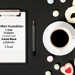 May include: A clipboard with a list of fonts to choose from: Arial, Caladea, Corbel Light, Arial Black, Gabriola, and Script. The clipboard is on a black surface with a cup of coffee, a pen, and meringue cookies.