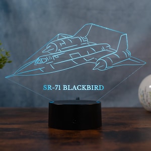 Puede incluir: Una luz nocturna negra con un avión SR-71 Blackbird. La luz presenta un contorno detallado del avión en color azul brillante, con el texto "SR-71 BLACKBIRD" debajo. La luz se encuentra sobre una base negra.